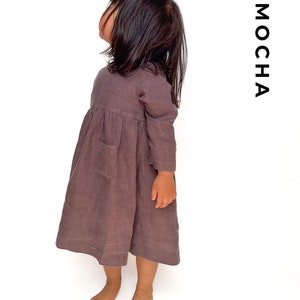 May include: A long-sleeved, brown linen dress with a pocket on the front. The dress is worn by a child and the word "MOCHA" is written vertically on the right side of the image.