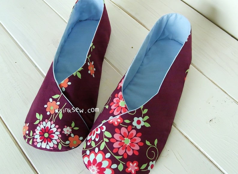 May include: A pair of burgundy fabric slippers with a floral pattern in pink, orange, and green. The interior is light blue, and the slippers have a wrap-around design. The slippers are designed for indoor use.
