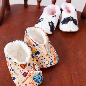May include: Two pairs of baby booties. One pair is white with black bear print and pink lining. The other pair is beige with a space-themed print and white lining. The booties are sitting on a wooden chair.