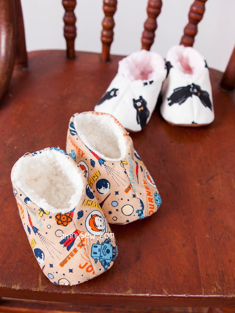 May include: Two pairs of baby booties. One pair is white with black bear print and pink lining. The other pair is beige with a space-themed print and white lining. The booties are sitting on a wooden chair.