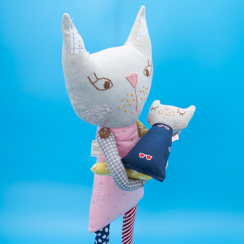 May include: A gray and white cat plush toy holding a smaller gray and white cat plush toy. The larger cat is wearing a pink dress with white stars and a checkered pattern on the sleeve. The smaller cat is wearing a blue vest with red sunglasses.