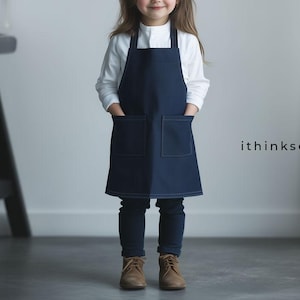 May include: A child wearing a white long-sleeved shirt and dark blue denim apron with two pockets. The apron has white stitching. The child is wearing dark blue pants and brown shoes. The text "ithinksew" is visible in the lower right corner of the image.