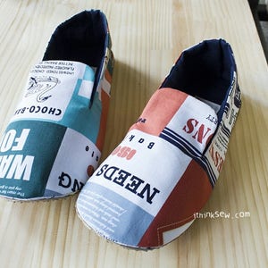 May include: A pair of blue and red fabric slippers with a patchwork design. The slippers have a variety of text printed on them, including "NEEDS", "BAK", "FO", and "CHOCO-BAN".