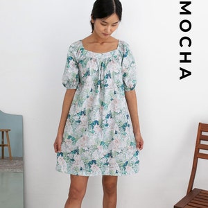May include: A short-sleeved dress with a square neckline and a floral pattern in shades of white, green, and blue. The word "MOCHA" is printed vertically in black on the right side of the image. The dress is knee-length.