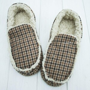 以下が含まれることがあります： 白のフリースの裏地が付いた、茶色と白の模様の入ったスリッパのペア。スリッパはシンプルなスリッポンデザインです。