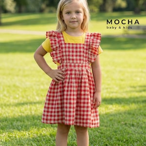 Young girl standing outdoors in a grassy park wearing a handmade red and white gingham ruffled baby pinafore dress over a yellow t-shirt. The digital sewing pattern creates a classic, versatile toddler jumper.