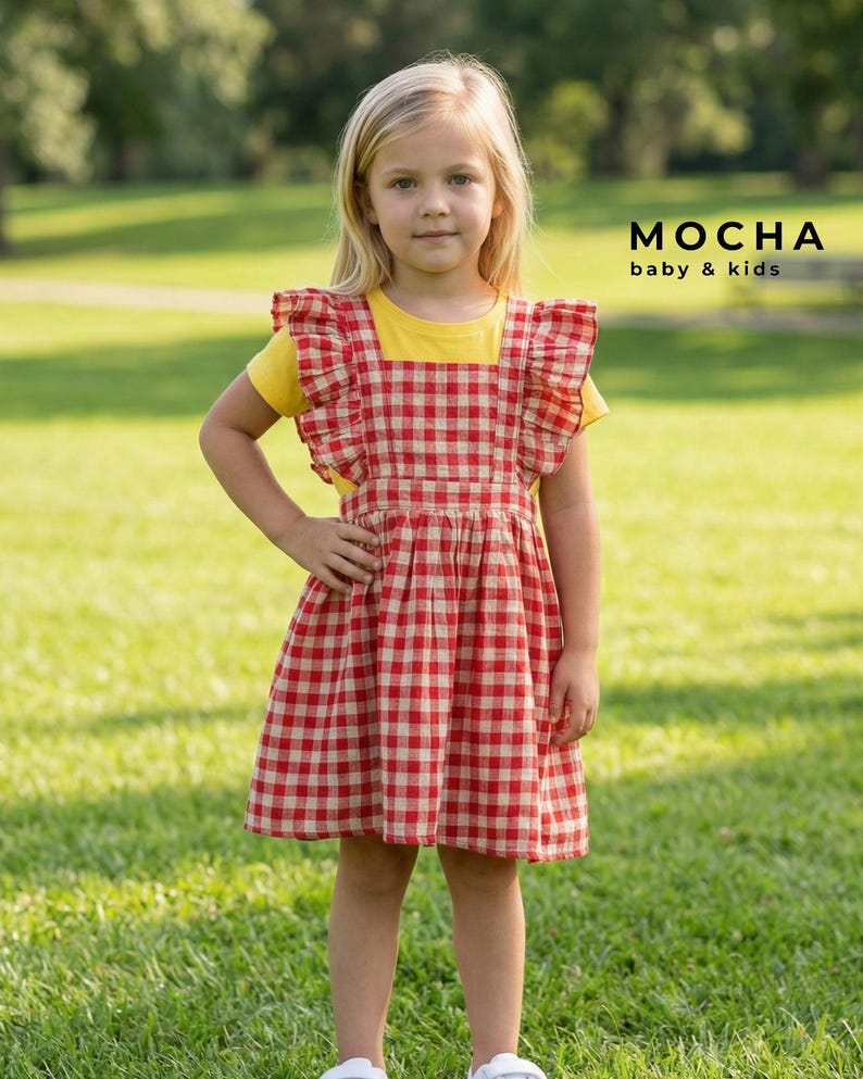 Young girl standing outdoors in a grassy park wearing a handmade red and white gingham ruffled baby pinafore dress over a yellow t-shirt. The digital sewing pattern creates a classic, versatile toddler jumper.