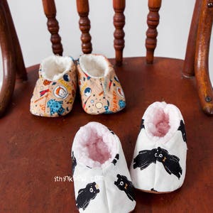 May include: Three pairs of baby booties. One pair is white with black bears, one pair is tan with a space theme, and one pair is tan with a rocket ship theme. The booties are sitting on a wooden chair.