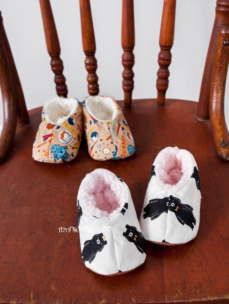 May include: Three pairs of baby booties. One pair is white with black bears, one pair is tan with a space theme, and one pair is tan with a rocket ship theme. The booties are sitting on a wooden chair.