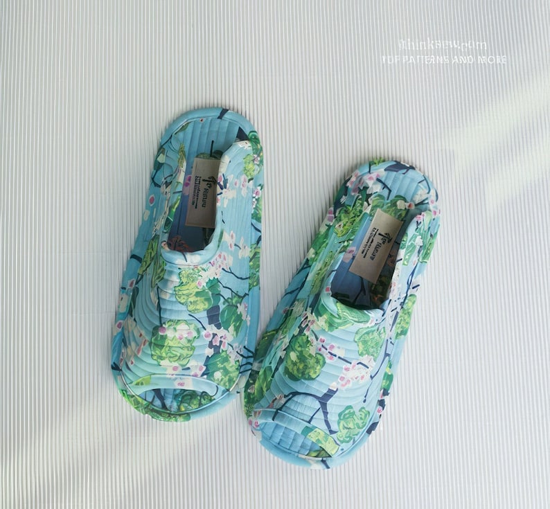 Op de afbeelding: Een paar blauwe en groene slippers met bloemenprint. De slippers hebben een gestikt ontwerp en zijn open.