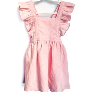 May include: A pink linen dress with ruffled straps and a gathered skirt. The dress is hanging on a white hanger.