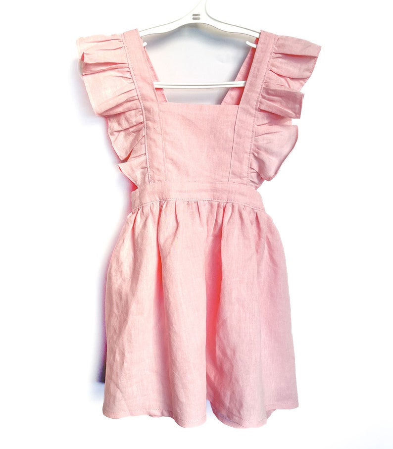 May include: A pink linen dress with ruffled straps and a gathered skirt. The dress is hanging on a white hanger.