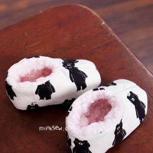 May include: A pair of white baby booties with a black bear print. The booties are lined with pink fleece and have a white background with black bears.