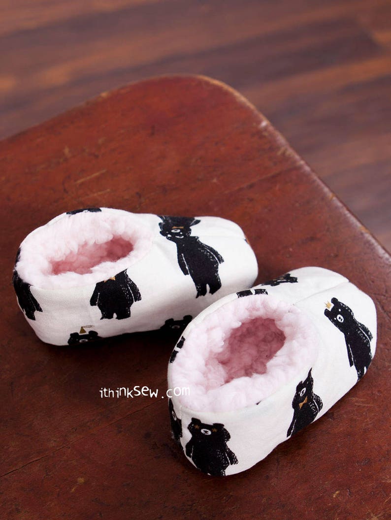 May include: A pair of white baby booties with a black bear print. The booties are lined with pink fleece and have a white background with black bears.