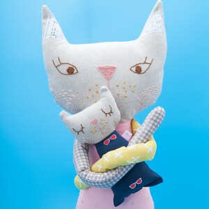 May include: A white plush cat doll with pink and brown embroidered details is holding a smaller gray plush cat doll with pink and brown embroidered details. The smaller cat doll is wearing a blue and yellow striped outfit with red sunglasses.