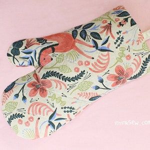 May include: A beige oven glove with a floral pattern featuring red, blue, and green flowers, leaves, and a stylised deer. The glove has a loop for hanging.