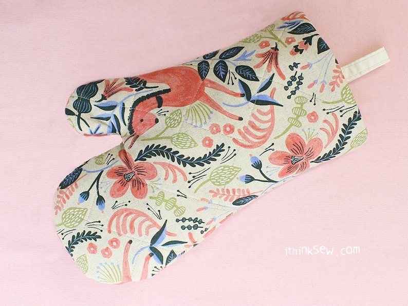 May include: A beige oven glove with a floral pattern featuring red, blue, and green flowers, leaves, and a stylised deer. The glove has a loop for hanging.
