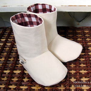 Könnte beinhalten: Ein Paar weiße Stoff-Stiefelüberzieher mit Gingham-Futter. Die Stiefelüberzieher liegen auf einer geflochtenen braunen und beigefarbenen Oberfläche.