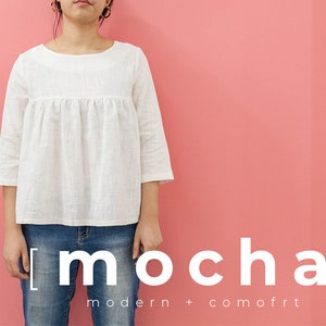 May include: A woman wearing a white, loose-fitting blouse with 3/4 sleeves and blue jeans. The text "[mocha] modern + comfort" is printed below the image.