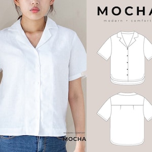 Puede incluir: Una camisa blanca de lino con cuello y mangas cortas. La camisa tiene un cierre de botones en la parte delantera y un ajuste holgado. La camisa se muestra en una modelo con jeans azules. El texto "MOCHA modern + comfort" se muestra en la parte superior de la imagen.