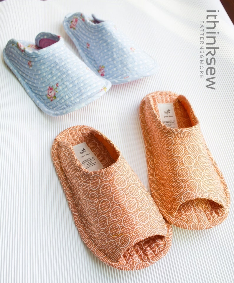 Op de afbeelding: Twee paar stoffen pantoffels. Een paar is lichtblauw met witte stippen en bloemenmotieven. Het andere paar is oranje met een wit cirkelpatroon. De pantoffels liggen op een wit oppervlak met een gestreept patroon.