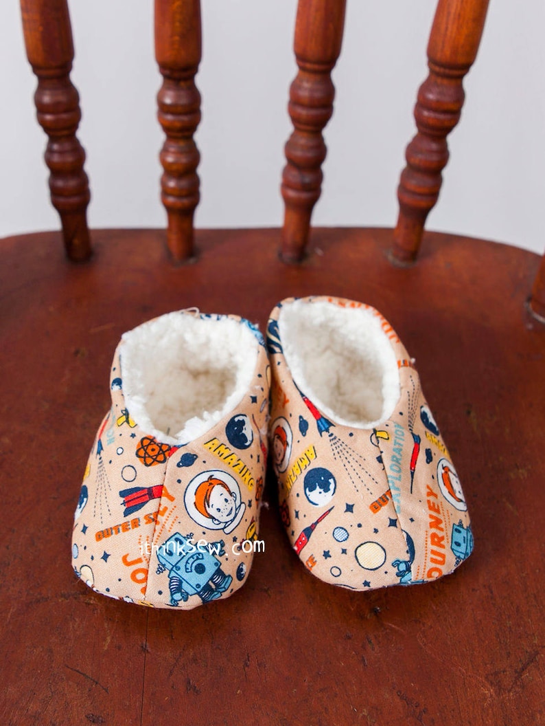May include: A pair of beige slipper booties with a space-themed print featuring rockets, planets, stars, and astronauts. The booties have a white fleece lining.