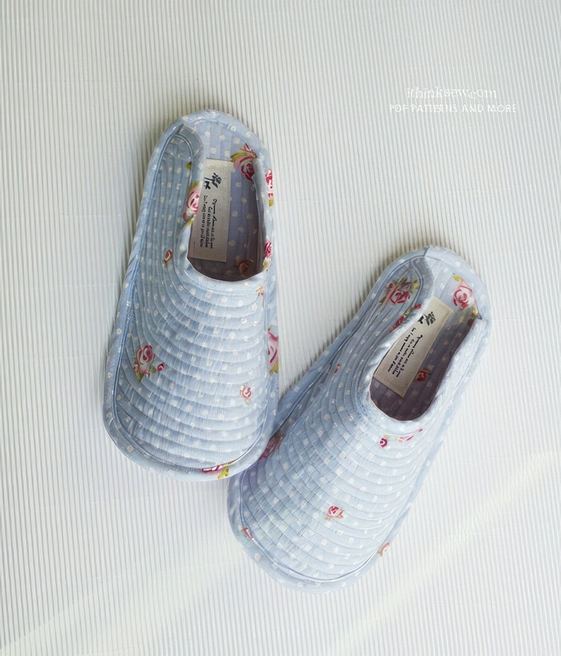 Op de afbeelding: Een paar blauwe, gewatteerde pantoffels met een bloemenprint. De pantoffels hebben een wit stippenpatroon en een wit label met tekst in het Japans.