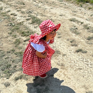May include: A young child wearing a red and white gingham dress and a red and white gingham sun hat. The child is standing on a dirt path with green grass on either side.