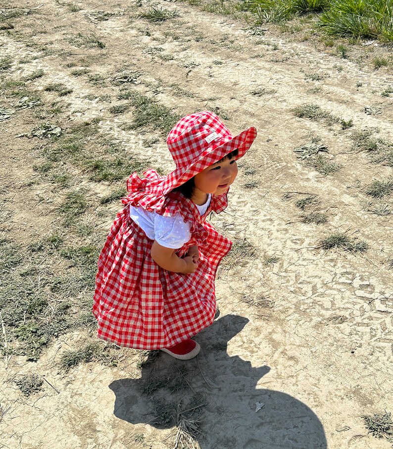 May include: A young child wearing a red and white gingham dress and a red and white gingham sun hat. The child is standing on a dirt path with green grass on either side.