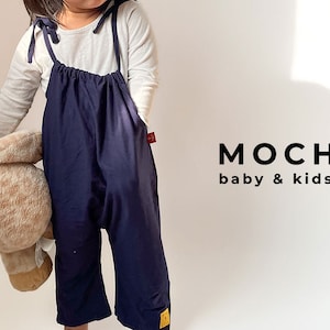 May include: A young child wearing a navy blue overall with a white long-sleeved shirt underneath. The overall has thin straps and a gathered waist. The child is holding a stuffed animal. The text "MOCHA baby & kids" is visible in the background.