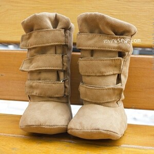 Puede incluir: Un par de botines de bebé de ante marrón con correas ajustables. Los botines están sobre una superficie de madera.