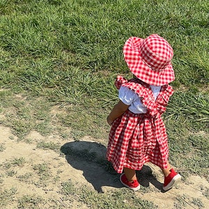 May include: A young child wearing a red and white gingham dress and matching sun hat stands on a dirt path. The child is facing away from the camera and looking to the right.