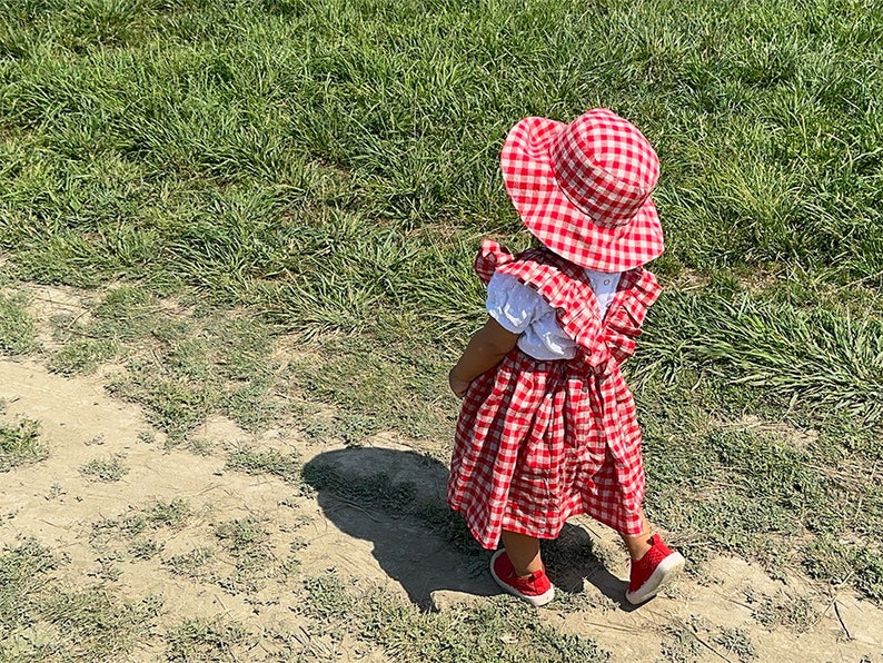 May include: A young child wearing a red and white gingham dress and matching sun hat stands on a dirt path. The child is facing away from the camera and looking to the right.
