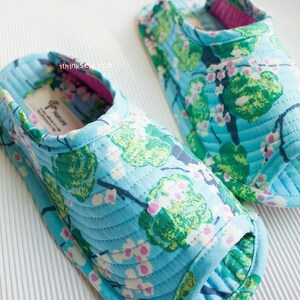 Op de afbeelding: Een paar blauwe, gewatteerde pantoffels met een bloemenprint van groene bladeren en roze bloemen. De pantoffels hebben een roze rand rond de opening.