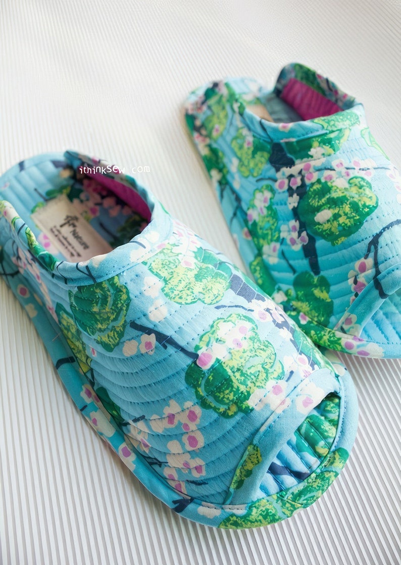 Op de afbeelding: Een paar blauwe, gewatteerde pantoffels met een bloemenprint van groene bladeren en roze bloemen. De pantoffels hebben een roze rand rond de opening.