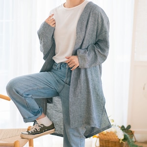 Może przedstawiać: Długi, jasnoniebieski kardigan z lnu o luźnym kroju. Kardigan ma nieco dłuższy tył i jest pokazany noszony z białym t-shirtem i niebieskimi dżinsami. Słowo "MOCHA" jest napisane pionowo czarnym tekstem po prawej stronie obrazu.