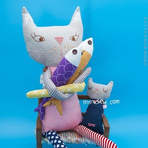 May include: Two plush cat toys, one larger than the other, are sitting on a wooden chair. The larger cat is holding two fish toys, one purple and one yellow. The smaller cat is wearing a blue and white striped outfit. The text "ithinksew.com" is visible on the smaller cat.