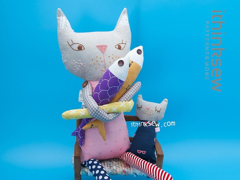 May include: Two plush cat toys, one larger than the other, are sitting on a wooden chair. The larger cat is holding two fish toys, one purple and one yellow. The smaller cat is wearing a blue and white striped outfit. The text "ithinksew.com" is visible on the smaller cat.