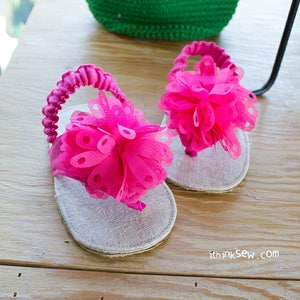 Könnte beinhalten: Ein Paar rosa und weiße Babysandalen mit einer großen rosa Blume an jedem Riemen. Die Sandalen liegen auf einer Holzoberfläche.
