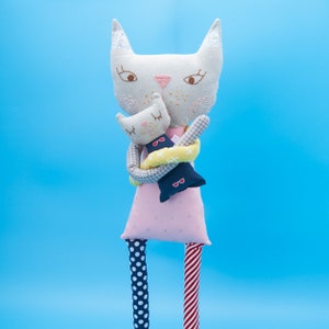 May include: A gray and white cat doll holding a smaller gray and white cat doll. The larger cat doll is wearing a pink dress with white polka dots and red and white striped stockings. The smaller cat doll is wearing a blue and white checkered dress with a yellow and white striped scarf.