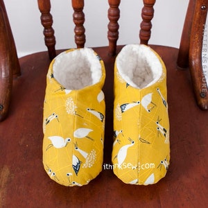 May include: A pair of yellow slippers with a white bird pattern. The slippers have a white fleece lining.