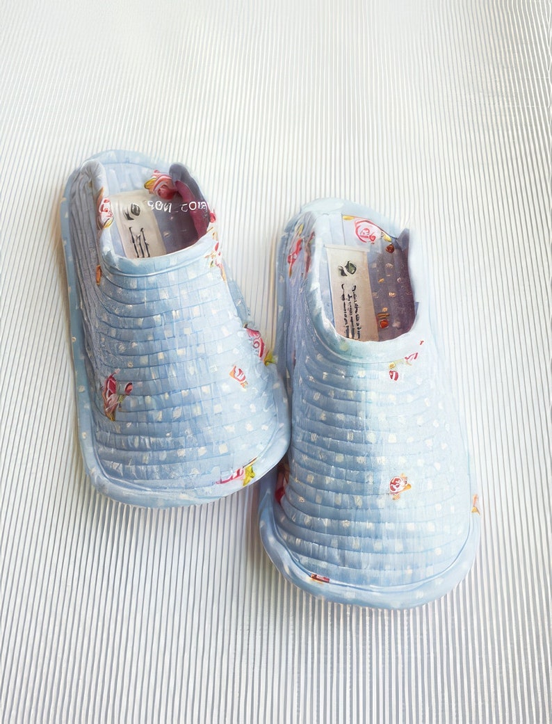 Op de afbeelding: Een paar lichtblauwe babyschoentjes met een wit stippenpatroon en kleine bloemenaccenten. De schoentjes hebben een gewatteerde textuur en zijn gemaakt van stof.