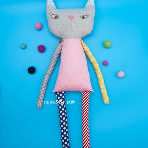 May include: A gray cat doll with pink, yellow, red, and blue patterned fabric arms and legs. The doll is wearing a pink dress with white polka dots. The doll is on a blue background with colorful balls scattered around it.
