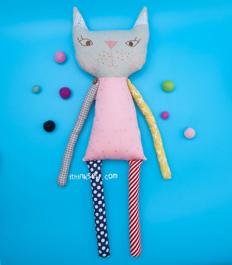 May include: A gray cat doll with pink, yellow, red, and blue patterned fabric arms and legs. The doll is wearing a pink dress with white polka dots. The doll is on a blue background with colorful balls scattered around it.