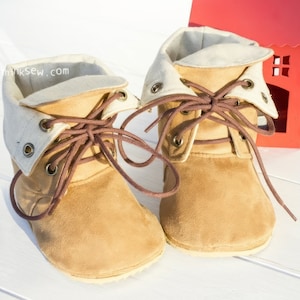 May include: A pair of brown suede baby booties with white fabric lining and brown laces. The booties are tied and sitting on a white wooden surface.