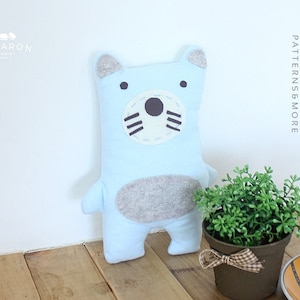 Może przedstawiać: Pluszowa zabawka w kolorze jasnoniebieskim w kształcie misia z szarymi uszami, szarym nosem i szarym brzuszkiem. Zabawka leży na drewnianej powierzchni z doniczką z rośliną i filiżanką kawy.