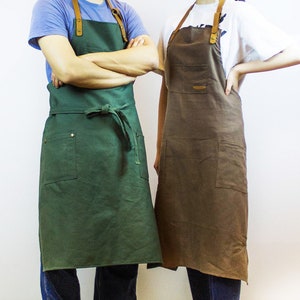 May include: Two aprons, one green and one brown, with leather straps and pockets. Both aprons have a simple design and are made of a durable fabric.