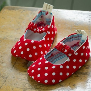Peut inclure: Une paire de chaussures de bébé rouges à pois blancs et une doublure rayée bleu et blanc. Les chaussures ont une sangle élastique blanche sur le dessus.