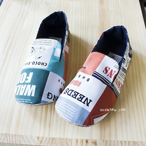 A pair of handmade men's slip-on fabric house shoes crafted from a sewing pattern, featuring a colorful retro typography print in blue, orange, and white, resting on a light wooden surface.