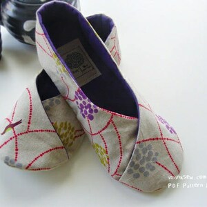 May include: A pair of beige fabric slippers with a floral pattern in purple, yellow, and gray, accented with red dotted lines. The slippers have a dark purple interior and a label. A teapot and teacup are in the background.